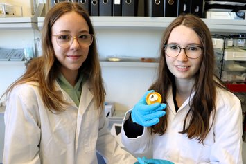 Preparation of carbon Two girls wearing white lab coats and blue protective gloves hold a prepared sample holder towards the camera.