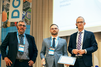 Georg Sachs Prize 2026 Konrad Kosiba Dr.-Ing. Konrad Kosiba stands on stage holding the Georg Sachs Prize 2026 medal and certificate, flanked by two representatives of the German Society for Materials Science.